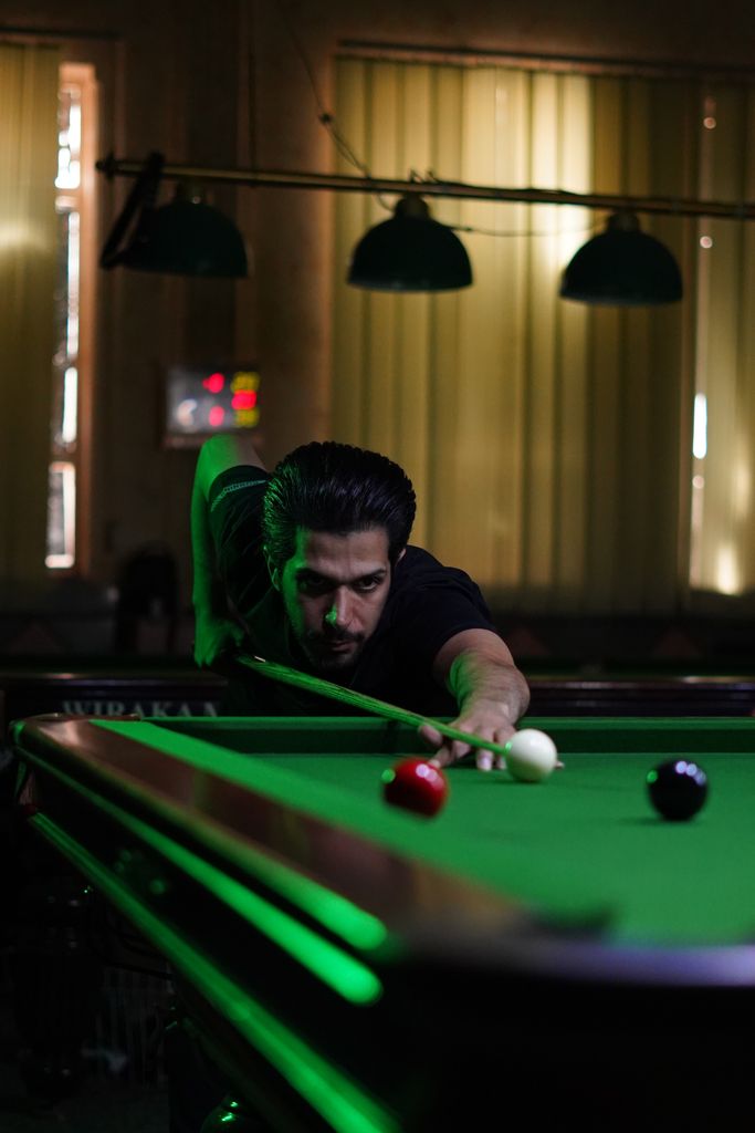 A man intently aiming to strike a snooker ball on a dimly lit snooker table, showcasing precise focus.